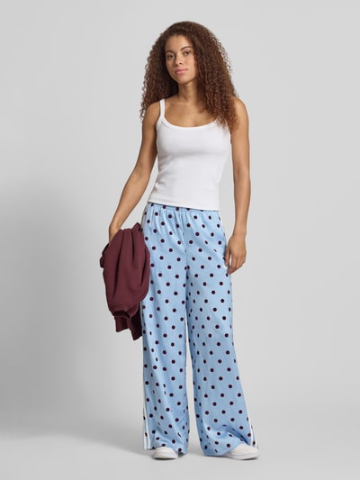 Frauenmodel trägt Loose Fit Sweatpants mit Polka Dots Modell 'CLESKY' von adidas Originals in Optik: Allover-Muster, Wide-Leg, Tunnelzug-Bundabschluss, Logo-Print, Streifen-Details, Materialverarbeitung: Satin, sanfte Blautöne mit dunklen Punkten, fließende glatte Textur, Ganzkörperansicht von vorne.