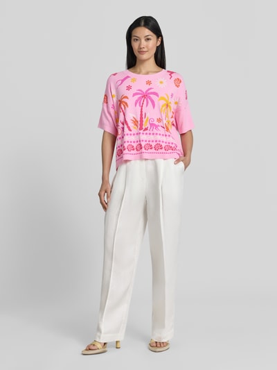 Modelka kobieca ma na sobie Strickshirt aus Viskose-Mix im Allover-Look od Milano Italy, róż z żółtym i pomarańczowym wzorem, luźny krój, obniżone ramiona, okrągły dekolt, długość tyłu w rozmiarze S: 50 cm, pełna sylwetka z przodu.