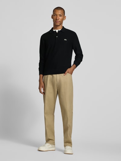 Mężczyzna model nosi Regular Fit Poloshirt aus reiner Baumwolle von Lacoste, czarna koszulka polo, beżowe spodnie, białe buty, Ganzkörperansicht von vorne, Herren Poloshirt von Lacoste.