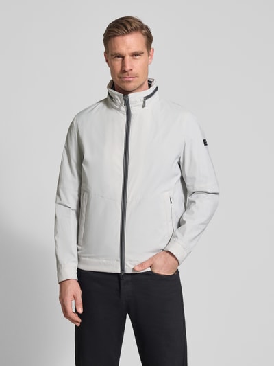Männermodel trägt Regular Fit Jacke mit Stehkragen von bugatti in lichtgrijs, zwart detail, Logodetail, steekzakken, tweewegritssluiting, capuchon, elastische mouwboord, ruglengte 68 cm, schouderbreedte 42 cm, borstwijdte 50 cm, mouwlengte 65 cm, ganzkörperansicht van voren.
