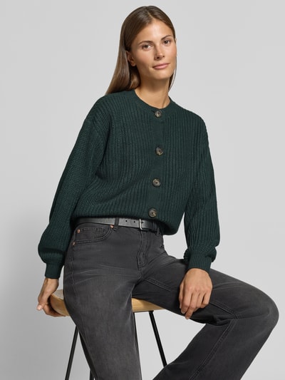 Frauenmodel trägt Loose Fit Strickjacke mit einreihiger Knopfleiste Modell 'LEA' von VERO MODA in Dunkelgrün, grobe Strickstruktur, Nahaufnahme Oberkörper.