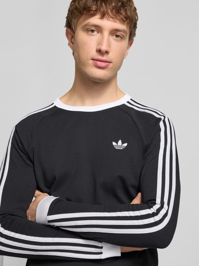 Männermodel trägt Longsleeve mit gerippten Abschlüssen von adidas Originals aus reiner Baumwolle in Schwarz mit weißen Details, Nahaufnahme Oberkörper.