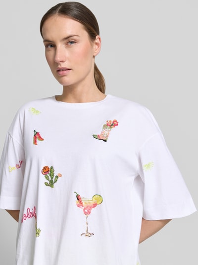 Modelka kobieca nosi T-Shirt mit Motiv-Stitchings od Marc Cain z czystej bawełny, krój loose fit, okrągły dekolt, biały z kolorowymi haftami i cekinami, długość pleców w rozmiarze 36: 55 cm, zbliżenie na górną część ciała z przodu.