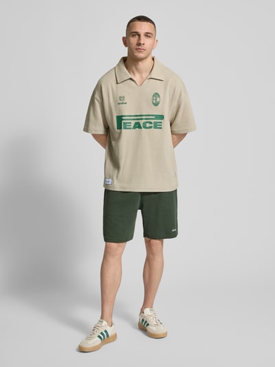 Männermodel trägt Shorts mit Logo-Print und Eingrifftaschen von ANOTHER COTTON LAB in Dunkelgrün, lockerer Schnitt, Ganzkörperansicht von vorne.
