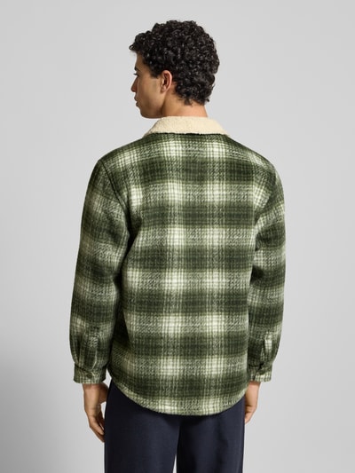 Męski model nosi Oversized Hemdjacke mit Allover-Muster od Tommy Jeans, efekt: wzór na całej powierzchni, krój oversized, wykładany kołnierz, listwa z zatrzaskami, naszywka z logo, długi rękaw, zakończenie rękawów na zatrzask, kieszenie boczne i na piersi, kolory: zielenie, beż, całość w kratę, tekstura miękka, Ganzkörperansicht Rückansicht.
