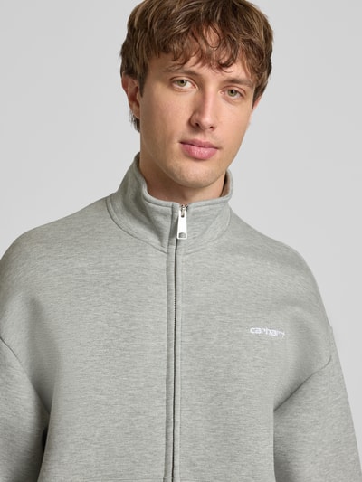 Männermodel trägt Sweatjacke mit Stehkragen von Carhartt Work In Progress in Hellgrau mit Reißverschluss und Label-Stitching, Nahaufnahme Oberkörper.