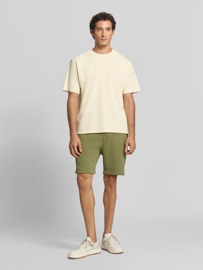 Männermodel trägt Shorts mit Label-Badge und elastischem Bund von Scotch & Soda in Olivgrün, reine Baumwolle, Regular Fit, Ganzkörperansicht von vorne.