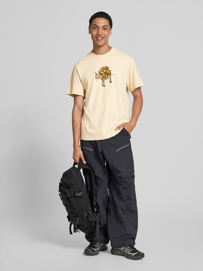 Männermodel draagt T-shirt met motiefprint model 'DONKEY TAXI' van The North Face, puur katoen, regular fit, ronde hals, heren, lichtbeige met gekleurde print, ruglengte bij maat M: 70 cm, vooraanzicht full body.