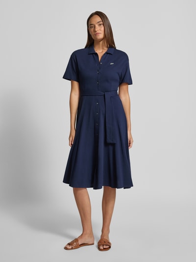 Frauenmodel trägt Tailliertes Freizeitkleid aus reiner Baumwolle von Lacoste in Dunkelblau, knielang, kurzarm, unifarben, Bindegürtel, Ganzkörperansicht von vorne.