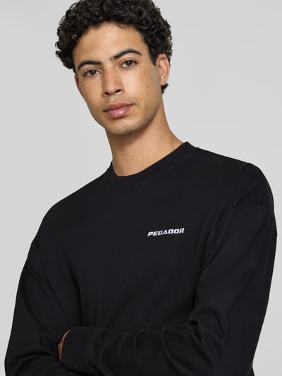 Männermodel trägt Oversized Sweatshirt mit Logo-Stitching Modell 'Rusk' von Pegador in Schwarz aus Reine Baumwolle, Nahaufnahme Oberkörper. Rückenlänge bei Größe M: 61 cm. Oversized-Fit.