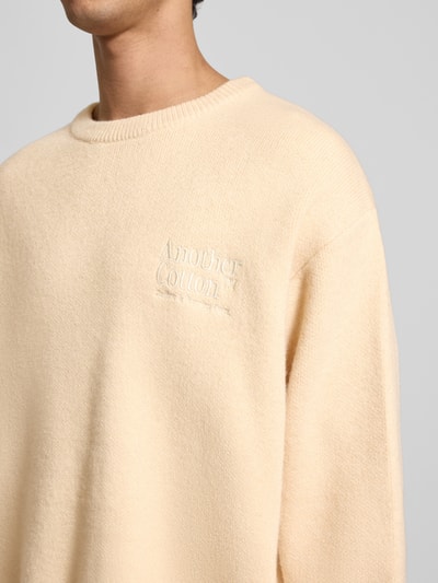 Männermodel trägt Strickpullover mit gerippten Abschlüssen von ANOTHER COTTON LAB in Beige, Rundhalsausschnitt, Label-Stitching, Nahaufnahme Oberkörper.