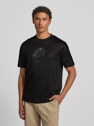 Männermodel trägt schwarzes T-Shirt mit Label-Detail von Emporio Armani aus Lyocell-Baumwoll-Mix, Regular-Fit, Rundhalsausschnitt, Ganzkörperansicht von vorne. Rückenlänge bei Größe M: 70 cm.