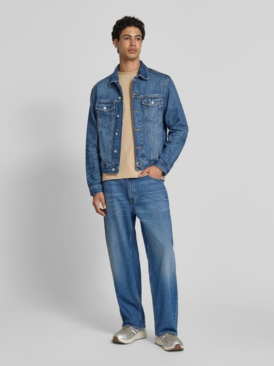Męski model nosi Regular Fit Jeansjacke aus reiner Baumwolle Modell 'RYAN' von Tommy Jeans, stylizowana na denim, wykończenie: splot twill, długi rękaw, wykładany kołnierz, listwa guzikowa, naszywka z logo, wytłaczane logo, pas na plecach, nity, mankiet, rzep, wpuszczane kieszenie boczne, kieszeń na piersi, bawełna; widok całej sylwetki z przodu
