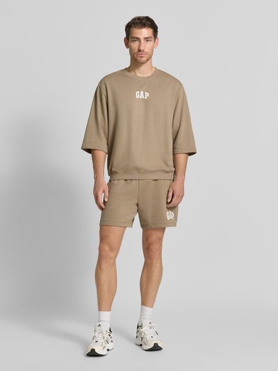 Männermodel trägt T-Shirt mit Rundhalsausschnitt und Logo-Print von GAP in beige aus katoenmix, loose fit, ribboorden, logoprint, ganzkörperansicht von vorne