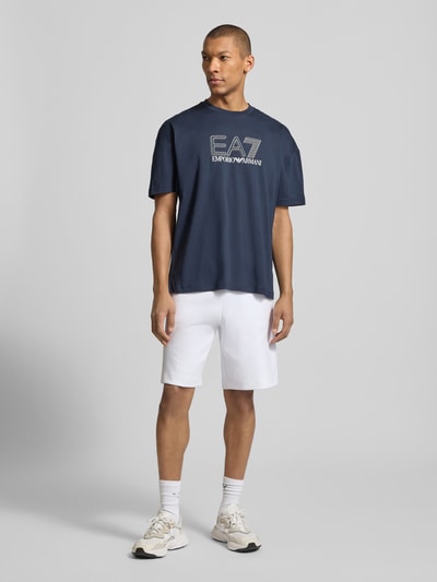Männermodel trägt T-Shirt mit Logo und Rundhalsausschnitt von EA7 Emporio Armani in Regular-Fit aus reiner Baumwolle, Ganzkörperansicht von vorne.