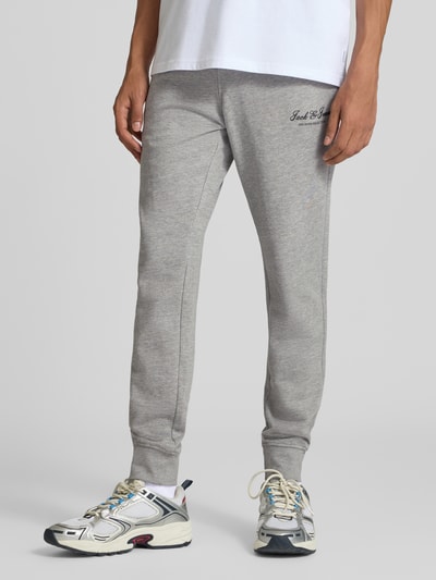 Männermodel trägt lichtgrijze sweatpants met labelprint Model 'GORDON' van JACK & JONES, los model, elastische band met tunnelkoord, zij-steekzakken, binnenbeenlengte bij maat M: 77 cm, detailopname onderlichaam, vooraanzicht