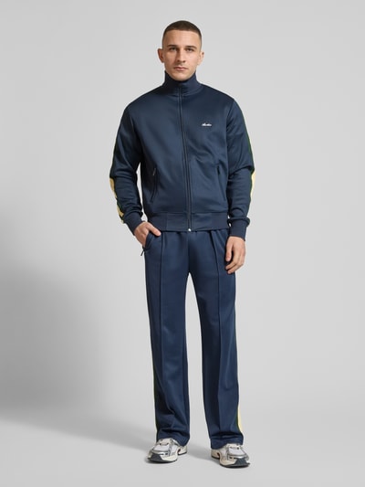 Männermodel trägt Track Jacket mit Stretch-Anteil Modell 'Everyday' von ANOTHER COTTON LAB, elastyczna mieszanka materiałów, prosty krój, stójka, zamek błyskawiczny, kieszenie zapinane na zamek błyskawiczny, elastyczne wykończenia, boczne lampasy, aplikacja z metką, długość tyłu w rozmiarze M: 63 cm, długość rękawów w rozmiarze M: 68 cm, granatowy z żółtymi detalami, gładka tekstura, Ganzkörperansicht von vorne