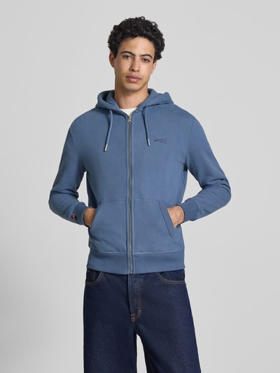 Männermodel trägt Sweatjacke mit Kapuze und Logo-Stitching von Superdry aus Baumwoll-Mix, Regular Fit, in Blau, Ganzkörperansicht von vorne.