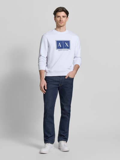 Męski model nosi Sweatshirt mit Logo-Print und Rundhalsausschnitt od ARMANI EXCHANGE, czysta bawełna, krój loose fit, biały z nadrukiem, dżinsy, buty sportowe, kolorystyka biała i granatowa, widok całej sylwetki z przodu.