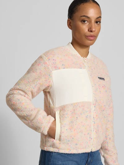 Modelka nosi Regular Fit Jacke mit Stehkragen od Columbia z polaru, jasnoróżowy kolor z kolorowymi akcentami, łatka z logo, kieszenie napoleonka i wpuszczane, zamek błyskawiczny, szerokość w rozmiarze S: 50 cm, długość tyłu w rozmiarze S: 49 cm, szerokość ramion w rozmiarze S: 44 cm, długość rękawa w rozmiarze S: 61 cm, krój regular fit, ujęcie: z przodu, z bliska.