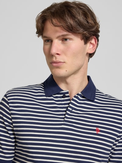 Męski model nosi Slim Fit Poloshirt mit Logo-Stitching von REVIEW z bawełny, kołnierzykiem polo, krótki rękaw, wyhaftowane logo, krój slim fit, długość tyłu w rozmiarze M: 67 cm, paski w granacie i bieli, tekstura dzianiny. Nahaufnahme górnej części ciała.