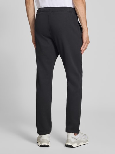 Mężczyzna nosi Sweatanzug mit Label-Details od Champion, kolor czarny, Mieszanka bawełny, prosty krój, Stójka, zamek błyskawiczny, detale z logo, wpuszczane kieszenie boczne, Elastyczny pas, Szerokość w rozmiarze M: 76,42 cm, Długość tyłu w rozmiarze M: 70 cm, Szerokość ramion w rozmiarze M: 51 cm, Długość rękawów w rozmiarze M: 69 cm, Długość wewnętrzna nogawki w rozmiarze M: 76 cm, czysta faktura, widok całej sylwetki od tyłu.