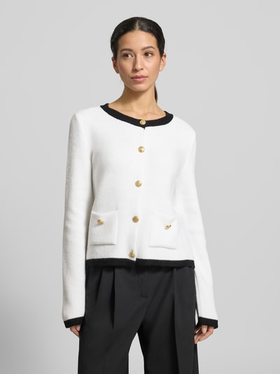 Vrouwenmodel draagt Slim Fit Jacke met Woll-Anteil in Strick-Optik van More & More, materiaalmix, tricotlook, ronde hals, opgestikte zakken, knoopsluiting, ruglengte maat 36: 53 cm, mouwlengte maat 36: 66 cm, kleur wit met zwarte accenten. Nahaufnahme bovenlichaam.