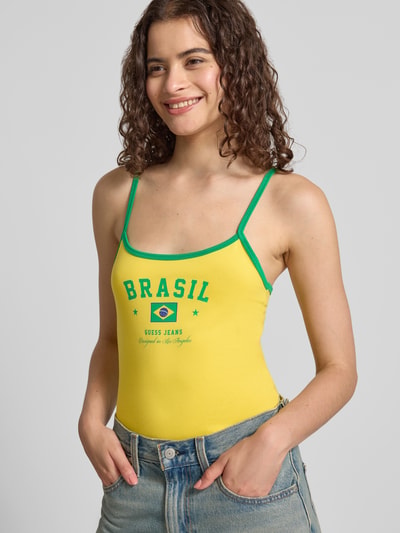 Frauenmodel trägt Slim Fit Top mit Statement-Print von GUESS in Gelb und Grün, elastischer Baumwoll-Mix, Nahaufnahme Oberkörper.