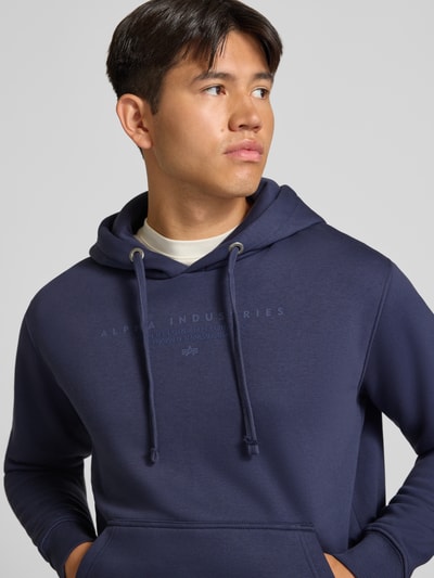 Männermodel trägt Relaxed Fit Hoodie aus Baumwoll-Mix mit Label-Detail von Alpha Industries in Dunkelblau, Nahaufnahme Oberkörper. Baumwoll-Mix, Relaxed Fit, Känguru-Tasche, Kapuze mit Tunnelzug, Label-Detail, elastische Abschlüsse.