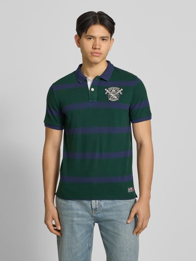 Männermodel trägt Slim Fit Poloshirt mit Label-Stitching und Knopfleiste von Superdry aus Reine Baumwolle, grün-blau gestreift, Slim-Fit Herren, Rückenlänge bei Größe M 70 cm, Ganzkörperansicht von vorne.