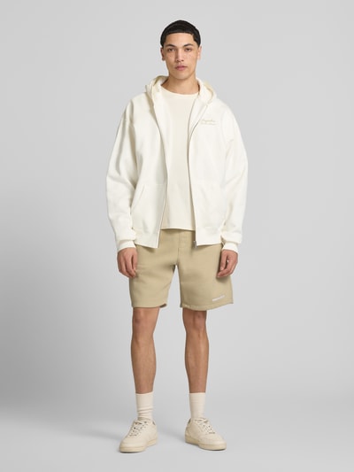 Männermodel trägt Shorts mit elastischem Bund und Logo-Stitching von Pegador aus Baumwoll-Mix, Loose-Fit, Eingrifftaschen, beige, Ganzkörperansicht von vorne.