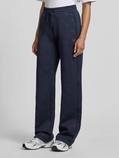Frauenmodel trägt dunkelblaue Wide-Fit Sweatpants mit Allover-Logo-Print Modell 'Calista' von Guess Activewear aus Elastischer-Material-Mix, mit Elastischer Bund und Tunnelzug, Innenbeinlänge 80 cm bei Größe S, Ganzkörperansicht von vorne.