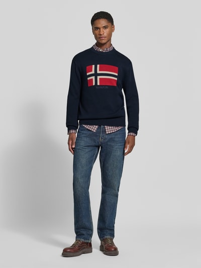Männermodel trägt gebreide pullover uit puur katoen Model 'MURA' van Napapijri, recht pasvorm, ronde hals, ribboorden, donkerblauw met rood-witte vlag op de borst, herenmode, jeans en leren schoenen, Ganzkörperansicht von vorne. Ruglengte maat M 67 cm, mouwlengte 70 cm.