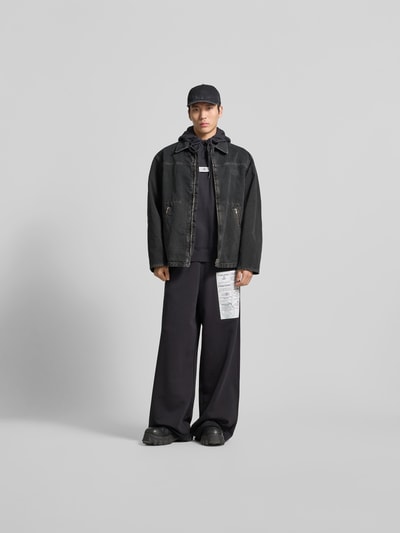 Männermodel trägt Jacke in Denim-Optik mit Umlegekragen von MM6 Maison Margiela in Schwarz, Reißverschlusstaschen, Relaxed Fit, Ganzkörperansicht von vorne.