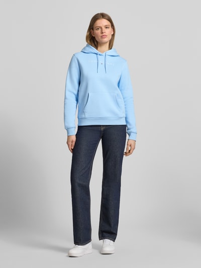 Frauenmodel trägt Regular Fit Hoodie aus Baumwoll-Mix von Tommy Jeans, unifarbenes Hellblau, Label Print, Sweatstruktur, Ganzkörperansicht von vorne.
