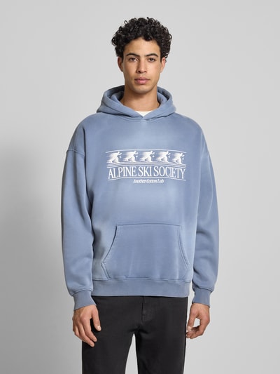 Männermodel trägt Oversized Hoodie mit Kapuze von ANOTHER COTTON LAB, Baumwoll-Mix, Label-Print, Känguru-Tasche, Ganzkörperansicht von vorne.