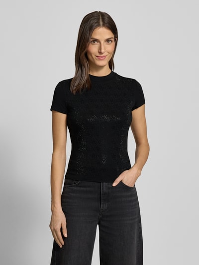 Kobiece modelka nosi Slim Fit T-Shirt mit Allover-Strasssteinbesatz od Guess, czarny, obszycie z kamieni stras na całej powierzchni, stójka, mieszanka bawełny i elastanu, detal z logo, długość tyłu 51 cm w rozmiarze S, widok całej sylwetki z przodu.