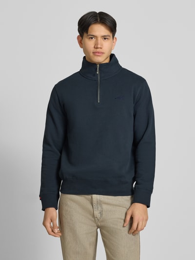 Męski model nosi Sweatshirt mit Reißverschluss und Label-Stitching od Superdry z mieszanki bawełny, krój regular fit, stójka, zamek błyskawiczny, wyhaftowane logo, długość tyłu w rozmiarze M: 67 cm, długość rękawów w rozmiarze M: 69 cm, widok całej sylwetki z przodu