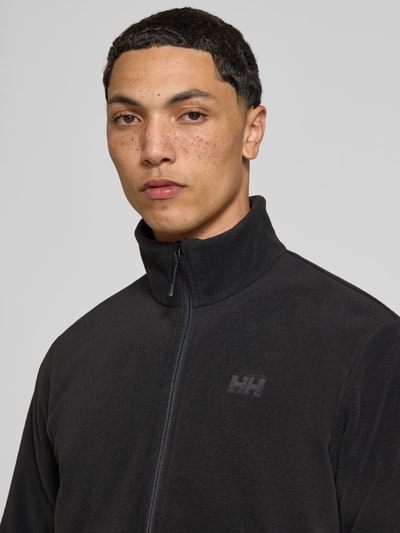 Männermodel trägt Regular Fit Fleecejacke mit Stehkragen Modell 'Daybreaker' von Helly Hansen in Schwarz, Nahaufnahme Oberkörper. Fleece, Reißverschluss, Eingrifftaschen mit Reißverschluss.