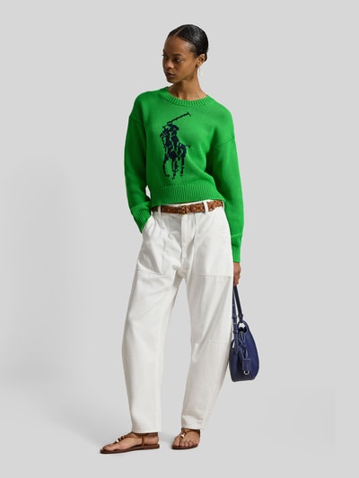 Frauenmodel trägt Regular Fit Strickpullover aus reiner Baumwolle von Polo Ralph Lauren, grün mit Two Tone Optik, Crew Neck, große Pony-Marke in Intarsia, Rippenabschlüsse, kombiniert zu weißer Hose, Ganzkörperansicht von vorne.
