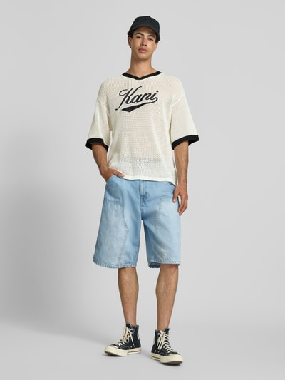 Männermodel trägt T-Shirt in Häkel-Optik mit V-Ausschnitt von KARL KANI aus Baumwoll-Mix, Loose-Fit, Label-Stitching, helle Farben, Ganzkörperansicht von vorne. Rückenlänge bei Größe M: 68 cm.