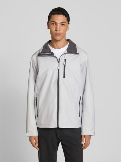 Männermodel trägt Regular Fit Jacke mit Stehkragen Modell 'Crew' von Helly Hansen, hellgrau mit schwarzem Reißverschluss, Ganzkörperansicht von vorne. Kontext: Herren Jacke, Stehkragen, Regular Fit, Modell 'Crew', Logo-Print, Napoleontasche, Eingrifftaschen mit Reißverschluss, Rückenlänge bei Größe M 73 cm, Schulterbreite 48 cm, Brustweite 56 cm, Ärmellänge 68 cm.