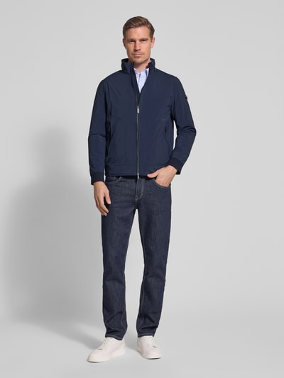 Męski model nosi Regular Fit Jacke mit Stehkragen od Bugatti, granatowa kurtka z zamkiem błyskawicznym, wpuszczane kieszenie, krój regular fit, zakończenia rękawów elastyczne, detal z logo, chowany kaptur, długość tyłu w rozmiarze 50: 65 cm, szerokość ramion: 47 cm, szerokość klatki piersiowej: 53 cm, długość rękawów: 63 cm, widok całej sylwetki z przodu.