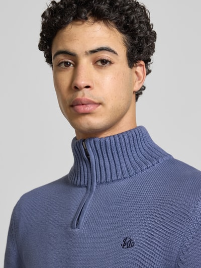 Männermodel trägt Slim Fit Pullover in Strick-Optik von Superdry in Blau mit Stehkragen, Reißverschluss, Logo-Stitching, Nahaufnahme Oberkörper.