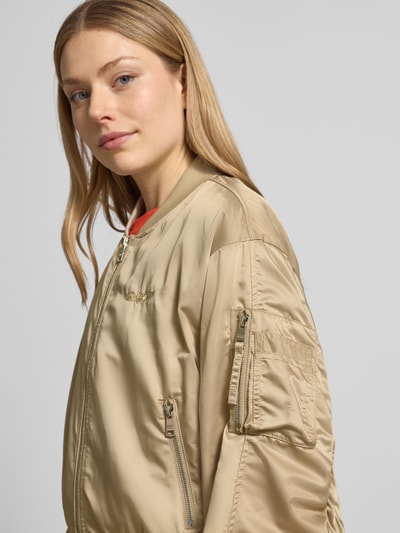 Vrouwenmodel draagt Bomberjacke met Reißverschlusstasche am Ärmel van Original Bombers in beige, glanzende textuur, los model, opstaande kraag, ritssluiting, ritszak op de mouw, gestikt logo, zij-steekzakken met drukknoopsluiting, labelpatch, elastisch bij de boorden, ruglengte maat S 49 cm, schouderbreedte 50 cm, borstwijdte 58 cm, mouwlengte 61 cm. Nahaufnahme bovenlichaam.