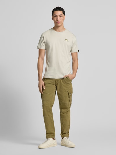Männermodel trägt T-Shirt aus Baumwolle von Alpha Industries in Beige mit Label-Details, gerader Schnitt, Rundhalsausschnitt, Ganzkörperansicht von vorne. Herren T-Shirt, Rückenlänge bei Größe M: 67 cm.