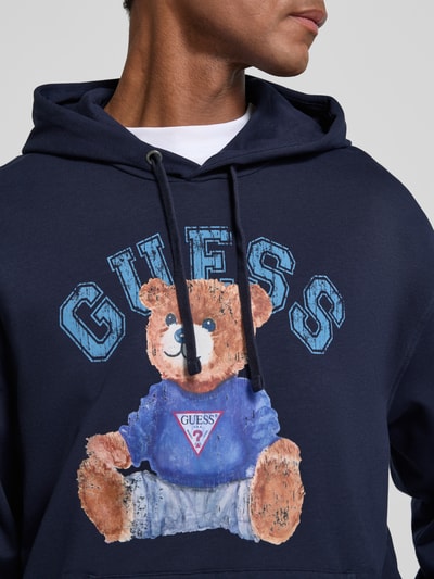 Mannelijk model draagt Hoodie mit Label-Print und Känguru-Tasche van GUESS in donkerblauw katoenmix met stretch, regular fit, labelprint en kangoeroezak, ruglengte maat M 71 cm, mouwlengte maat M 64 cm, detailopname bovenlichaam.