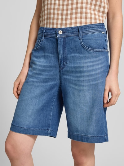 Frauenmodel trägt Regular Fit Jeansshorts aus Baumwoll-Mix Modell 'MAINE B' von Brax in Denim-Optik mit Label-Patch, 5-Pocket-Design, Knopf und verdecktem Reißverschluss, Gürtelschlaufen; mittleres Blau, leicht strukturierte Textur, Ganzkörperansicht von vorne.