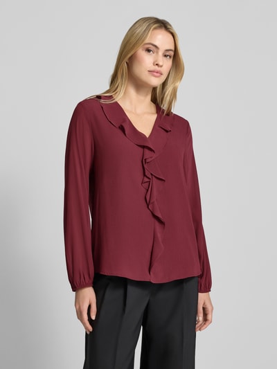 Modelka nosi Langarmbluse mit Rüschenvolant von S. OLIVER BLACK LABEL, kolor jednolity, dekolt w serek, długie rękawy z elastycznym mankietem, aplikacja falbany, dół z falbaną, widok całej sylwetki z przodu.