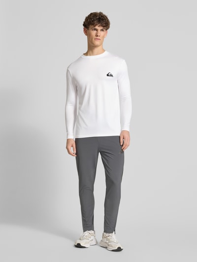 Männermodel trägt weißes Langarmshirt mit Sonnenschutz Modell 'EVERYDAY' von Quiksilver, körpernaher Schnitt, Ganzkörperansicht von vorne.